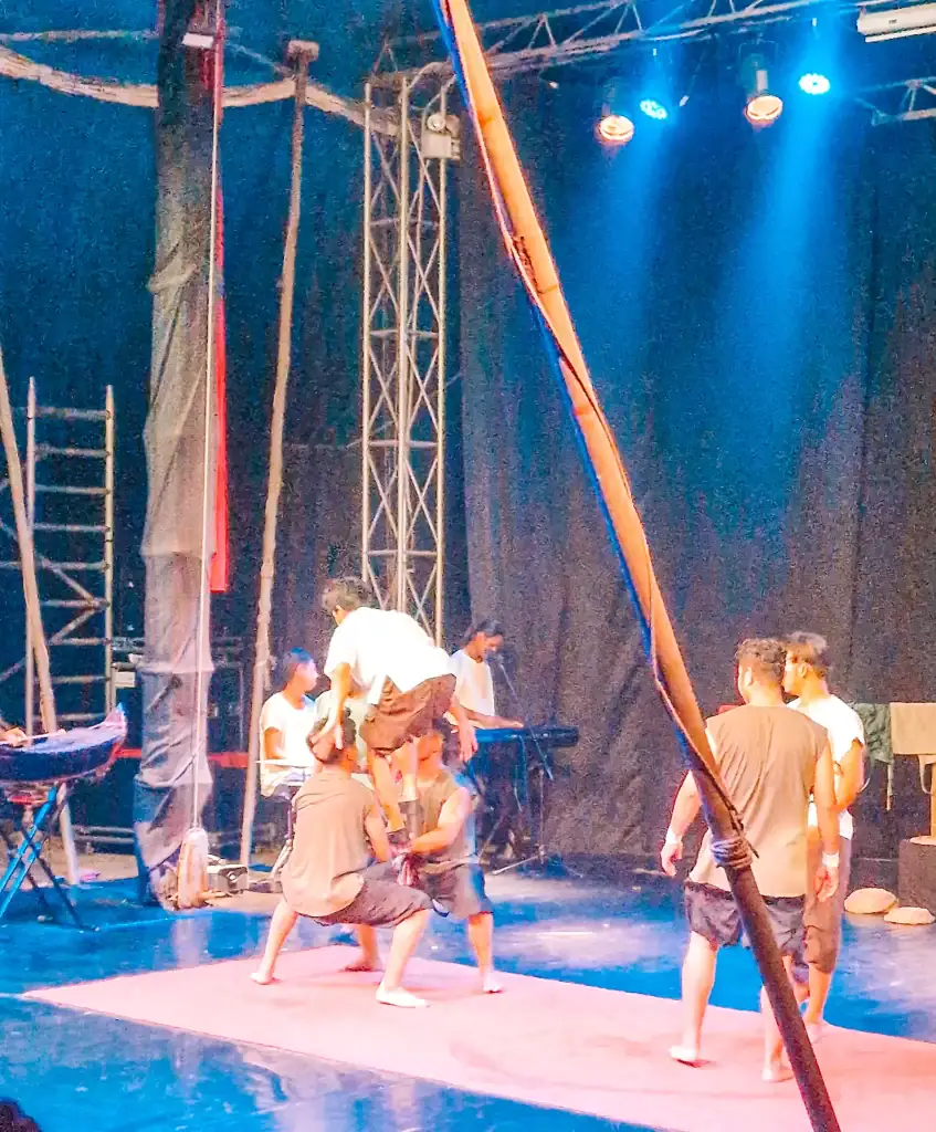 Performers mid-act during the Phare Cambodian Circus in Battambang, balancing and building a human pyramid on stage with blue lights overhead.