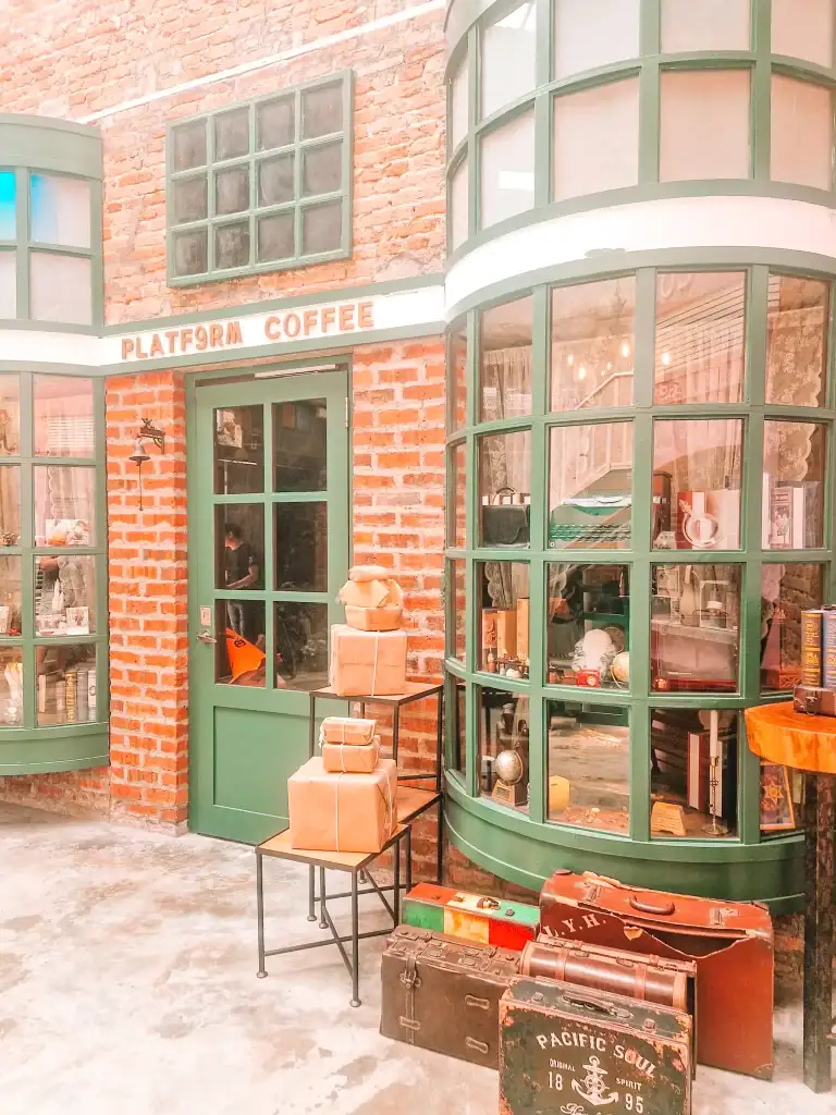 Cosy, vintage-themed café with green window frames and a brick façade, with parcels and suitcases stacked outside under the sign “PLATF9RM COFFEE,” blending industrial charm and travel nostalgia.