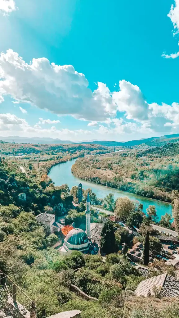 View from Počitelj Fortress overlooking a turquoise river winding through lush green hills, with a mosque and domed rooftops nestled in the foreground. A scenic must-see when exploring things to do near Mostar.