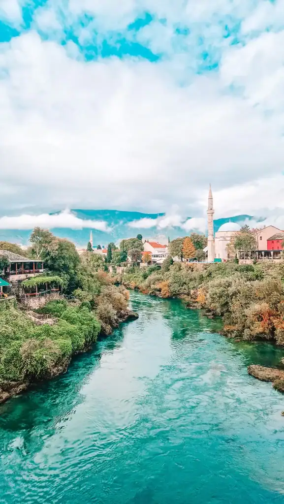 The emerald Neretva River flows through the center of Mostar, framed by leafy trees, colorful buildings, and a tall minaret. A peaceful riverside scene showcasing the town’s Ottoman architecture.