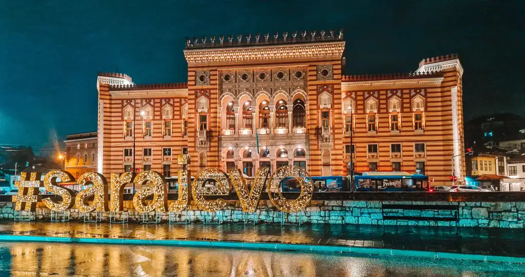 Nighttime view of Sarajevo City Hall lit up in golden tones, with a large illuminated #sarajevo sign in front. The wet pavement reflects the lights, adding a magical feel to this iconic building and must-see photo spot.