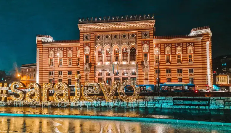 Nighttime view of Sarajevo City Hall lit up in golden tones, with a large illuminated #sarajevo sign in front. The wet pavement reflects the lights, adding a magical feel to this iconic building and must-see photo spot.