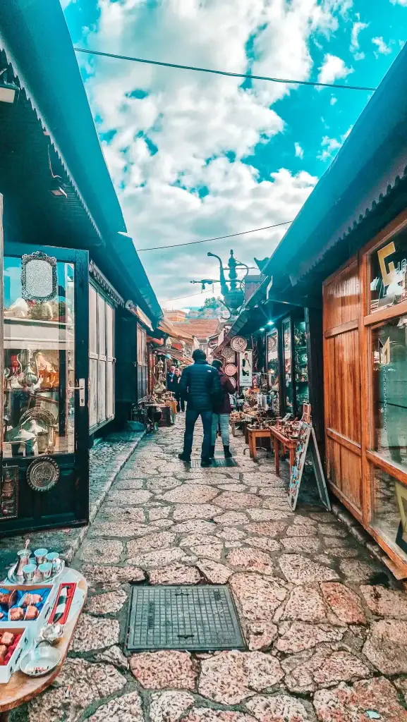Narrow cobblestone lane in Sarajevo’s Baščaršija lined with shops selling copperware and souvenirs, with people browsing under a bright sky. A top pick for things to do in Sarajevo for lovers of local crafts and market culture.