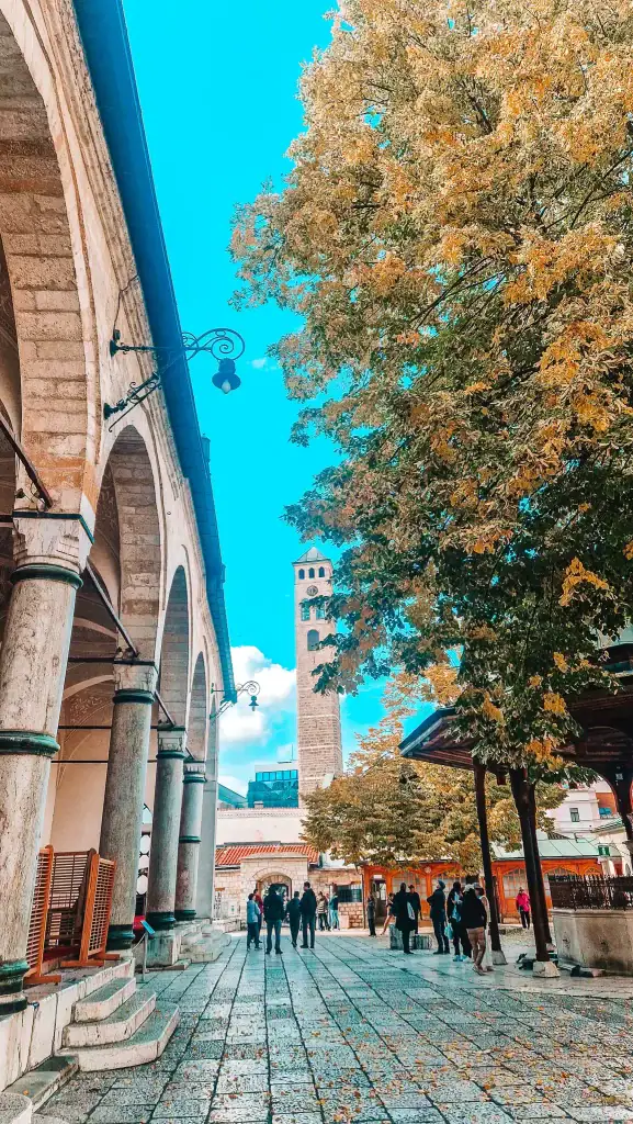 People walk through the courtyard of Gazi Husrev-beg Mosque, with a view of its clock tower and golden autumn trees under a clear sky. This peaceful and historic spot is a key site in Sarajevo’s old town.