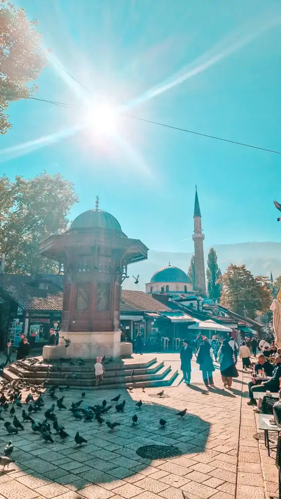 Sunlit Baščaršija square with the wooden Sebilj fountain at the centre, surrounded by pigeons, people, and historic architecture. The minaret in the background and the lively atmosphere make it one of the top things to do in Sarajevo.