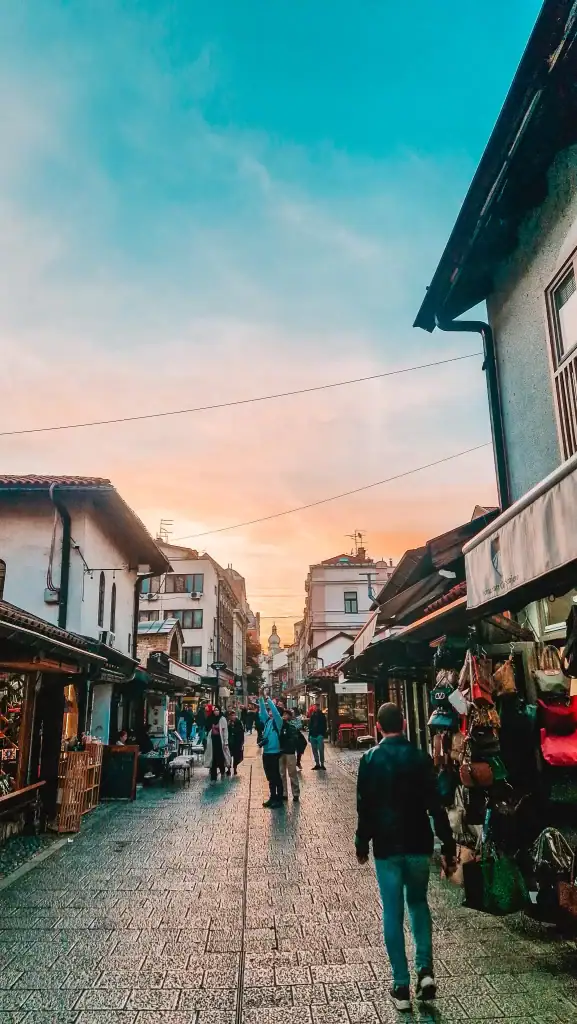 Evening street scene in Sarajevo’s old town with shoppers, souvenir stalls, and restaurants lining both sides of a cobbled path. The golden-hour sky casts a soft glow, adding charm to this must-visit neighbourhood.