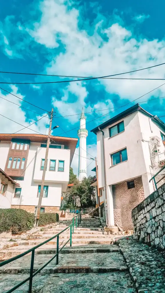 Stone steps lead uphill between traditional Bosnian homes with a white minaret rising in the background, framed by power lines and a vibrant blue sky. A peaceful residential view that hints at the city’s layered urban character.