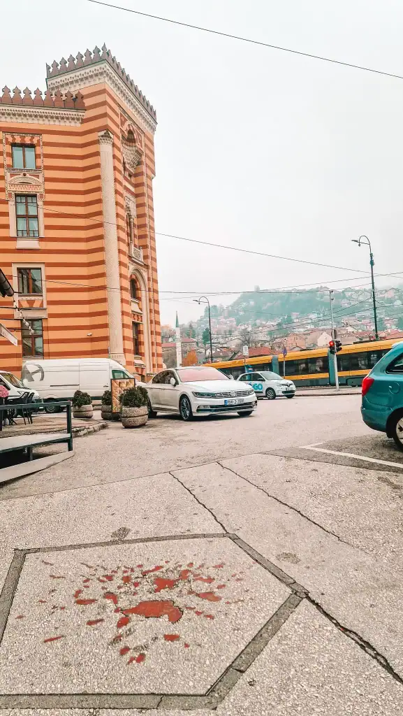 Red resin-filled Sarajevo Rose embedded in the pavement in front of the striking red-and-white striped Sarajevo City Hall, with trams and city buildings in the background. A powerful visual reminder of wartime scars in one of the city’s most visited areas.
