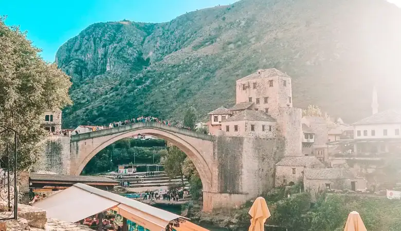 Golden sunlight beams over Stari Most and a riverside café packed with diners enjoying sunset views. A romantic and iconic experience in Mostar.