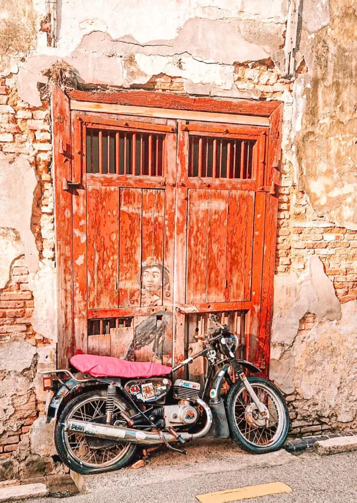 A piece of Penang’s famous street art showing a painted boy peeking through a red wooden door behind a real parked motorbike, merging mural and object into a 3D artwork.