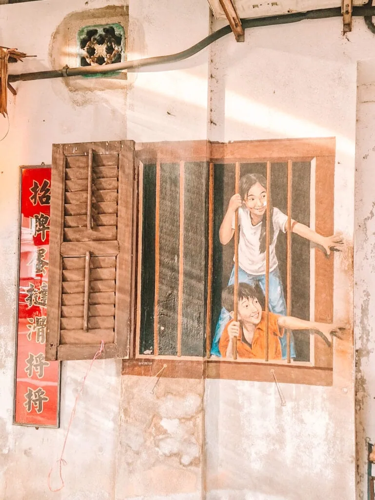 Mural of two children painted to look as though they are climbing through barred windows, blending playfully with the building’s actual architectural features.