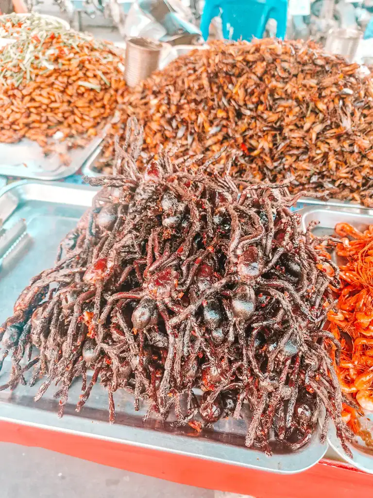 Close-up of a heaped metal tray of deep-fried tarantulas at a local Cambodian market, surrounded by other fried insects like crickets and beetles.