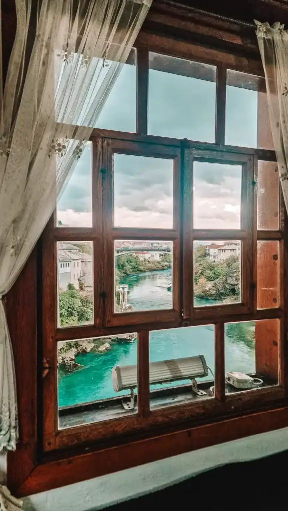 Looking through a wooden window with lace curtains at a view of the turquoise river and stone houses of Mostar. A tranquil perspective capturing the charm of local architecture.