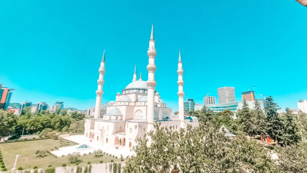 The Grand Mosque of Tirana stands tall with its four minarets and large domes, surrounded by modern high-rise buildings and lush city greenery.