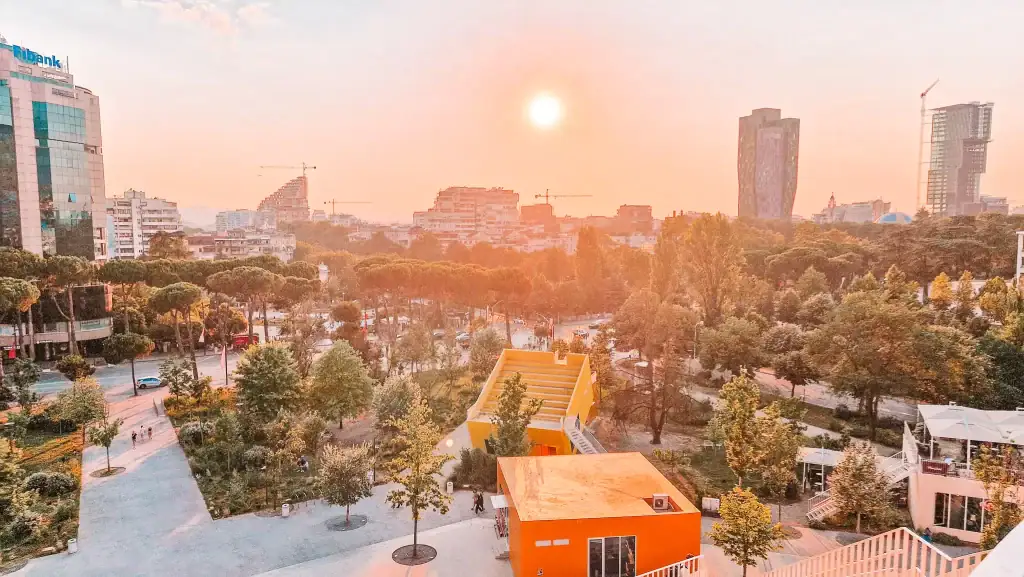 A warm sunset bathes downtown Tirana in golden light, with modern architecture, tree-lined streets, and the vibrant yellow staircase of Reja Art Installation in the foreground.
