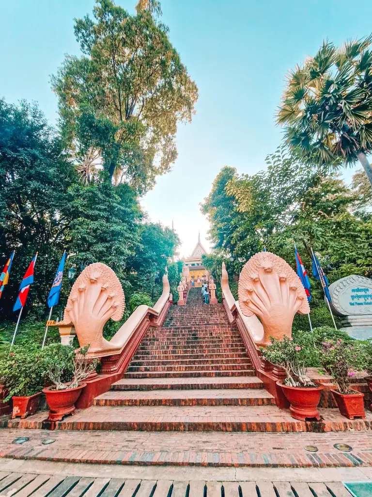 The grand red-brick staircase leading to Wat Phnom in Phnom Penh, lined with pink naga serpent statues and colourful flags, framed by tall trees under a blue sky.