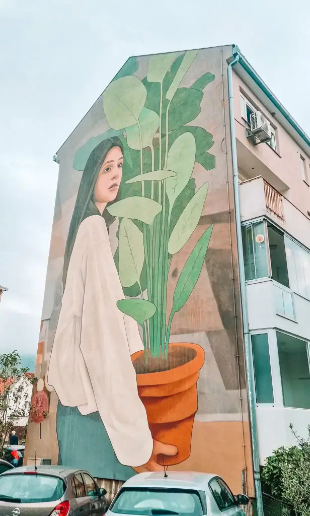 Large mural of a woman in a white blouse holding a potted plant, painted across the side of a residential building in Mostar. A striking example of the city’s contemporary street art scene.