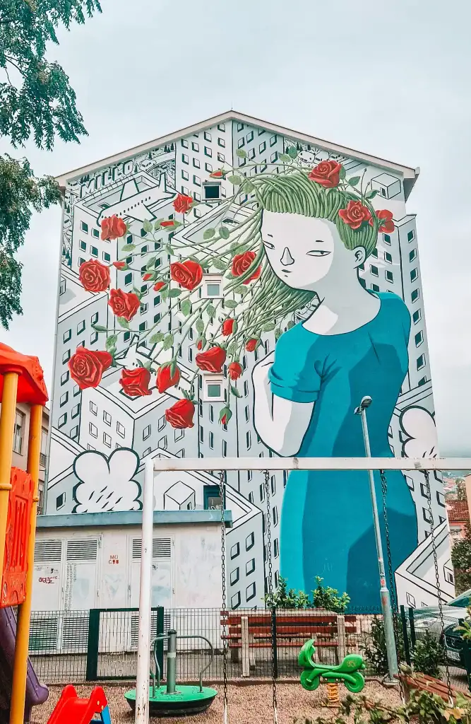 Whimsical mural of a girl in a blue dress with roses and vines flowing from her hair, set against a backdrop of cartoonish buildings. The painting covers the entire building façade above a playground in Mostar.