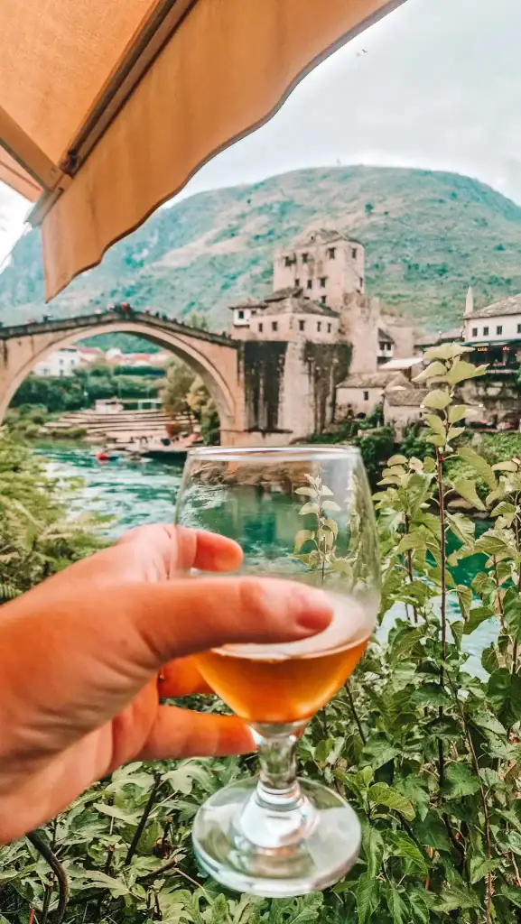 Hand holding a glass of beer in the foreground with the famous Stari Most bridge blurred in the background. A scenic and relaxing way to experience Mostar’s old town from a riverside café.