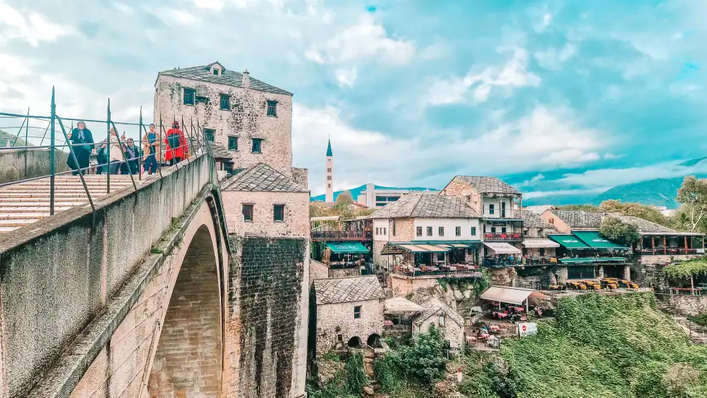Side view of Stari Most with tourists crossing the arched bridge and historic buildings stacked along the cliffs. An essential photo stop when listing top things to do in Mostar.