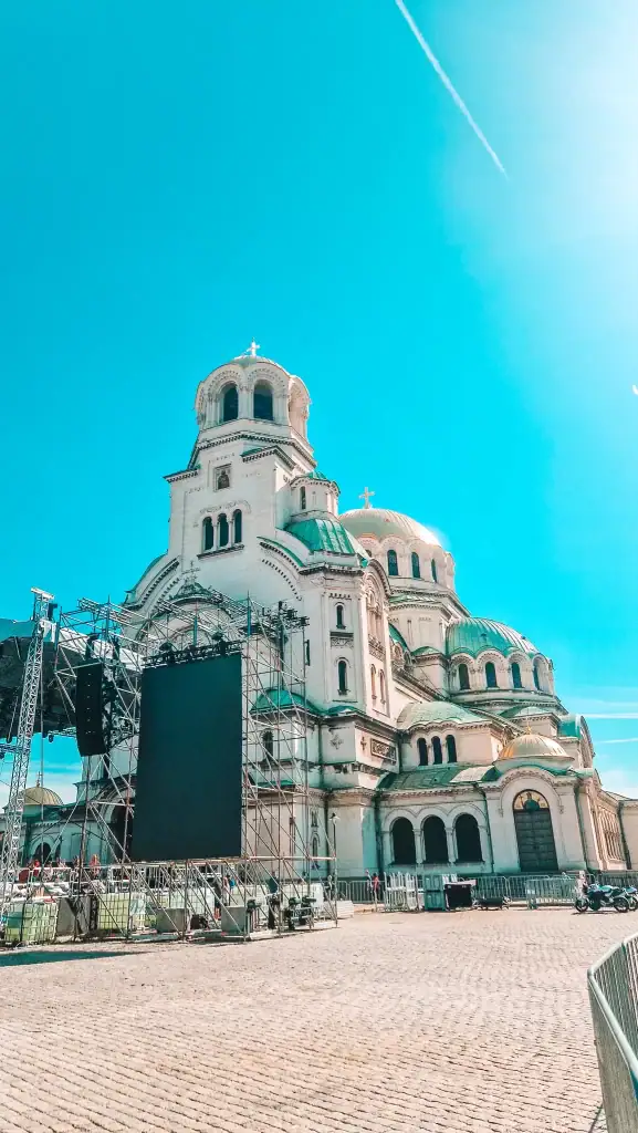 The Alexander Nevsky Cathedral in Sofia, Bulgaria, stands tall under a vibrant blue sky, with temporary stage scaffolding set up in the foreground. The cathedral’s gold and green domes gleam in the spring sunshine.