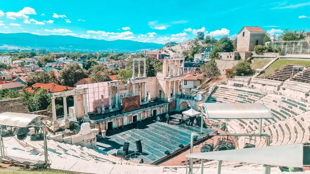 The ancient Roman Theatre of Plovdiv, Bulgaria, with white stone seating curving around a central stage and panoramic views of the city and surrounding mountains on a clear spring day.