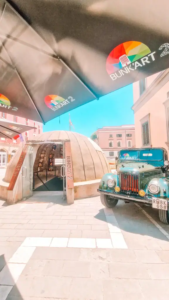 The entrance to Bunk’Art 2 in Tirana, Albania, features a domed concrete bunker with glass doors, a vintage military jeep parked beside it, and large parasols marked with the Bunk’Art logo.