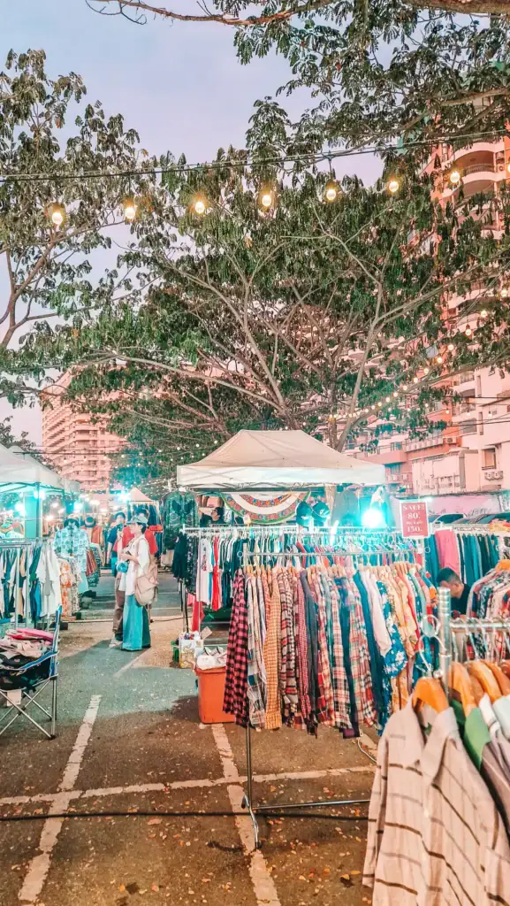 Colorful racks of secondhand clothes at a lively outdoor night market in Thailand with shoppers browsing under hanging string lights.