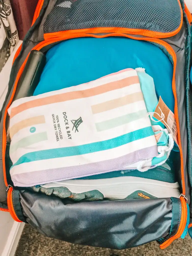 Close-up of an open Cotopaxi backpack packed with a striped Dock & Bay towel, hiking shoes, and travel essentials.