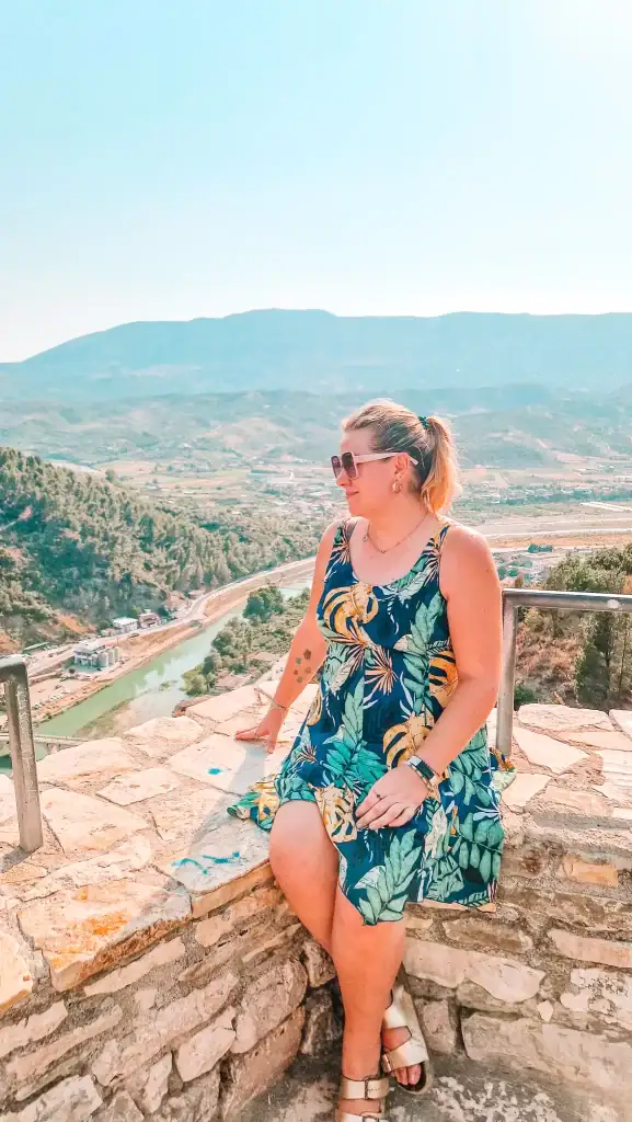 Em sits on a stone wall overlooking the Osum River in Berat, Albania, wearing a tropical dress and sunglasses, with rolling hills and a winding road visible in the bright spring landscape behind her.