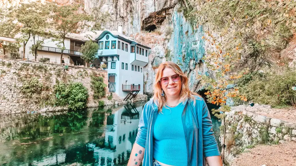Em stands in front of the serene turquoise water and cliffside Tekke of Blagaj in Bosnia and Herzegovina, smiling in a blue outfit with rocky terrain and a cave opening in the background.