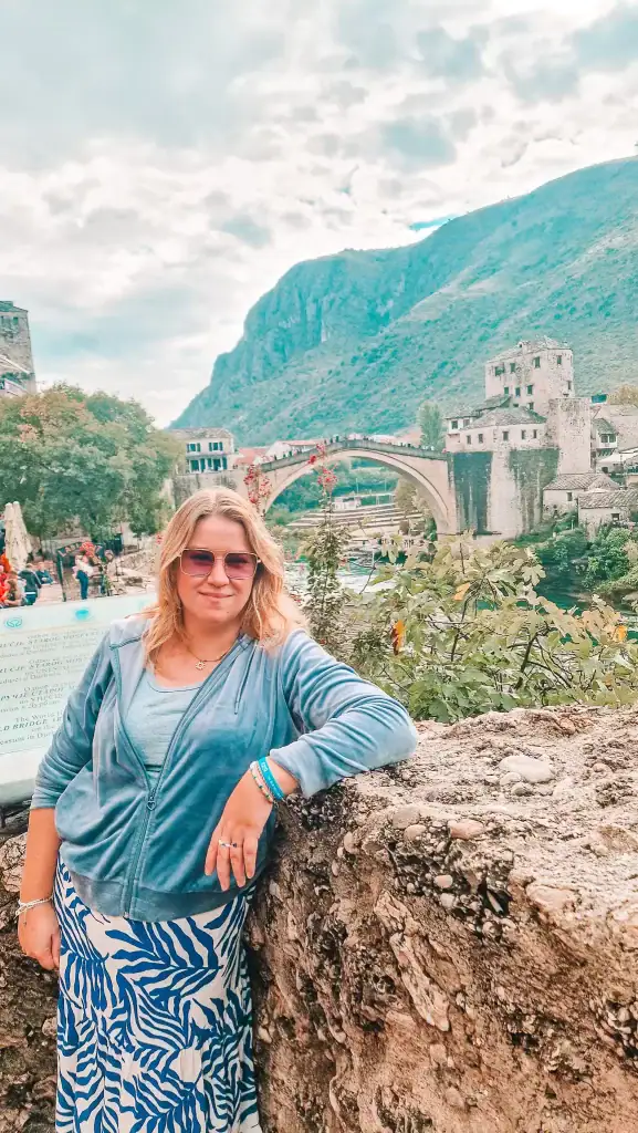 Em leans against a stone wall in front of the iconic Stari Most bridge in Mostar, Bosnia and Herzegovina, wearing a blue jacket and skirt as cloudy skies hover above the scenic old town and riverside.