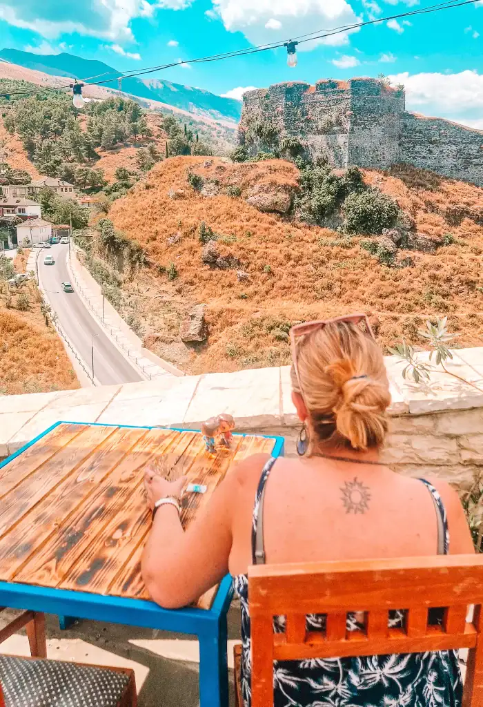 Em sits at a cafe table on a terrace in Gjirokastër, Albania, looking out at a winding road and stone fortress ruins on a golden hillside, with toy figures placed in front of her.