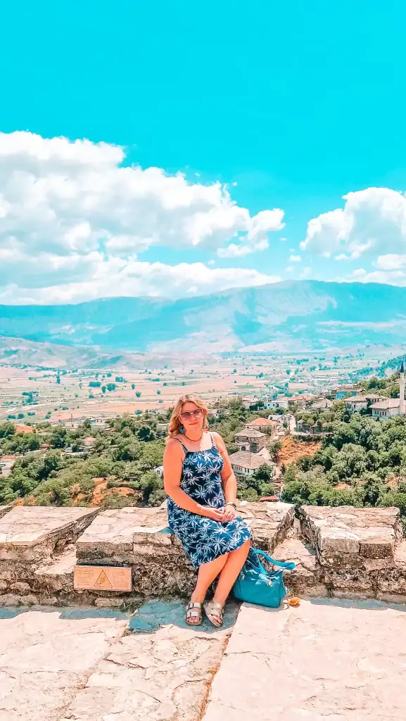 Em sits on a stone wall at Gjirokastër Castle in Albania, wearing a blue floral dress and sandals, with a sweeping springtime view of the valley, mountains, and traditional village below.