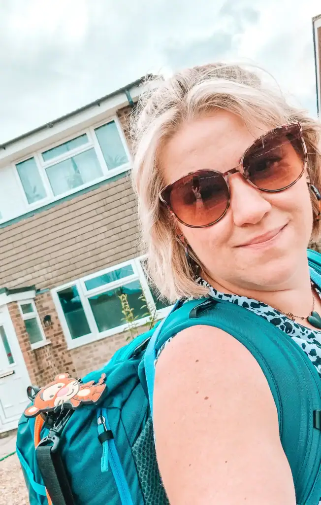 Em wearing sunglasses and a fully packed Cotopaxi backpack, standing outside a suburban home before departing on a trip.