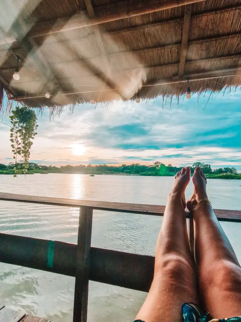 Em's legs stretched out toward a calm river while relaxing under a thatched roof in a wooden hut hotel in Laos with no bedside plug socket.