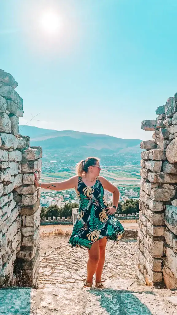 Framed by two stone walls, Em stands in a playful pose at Gjirokastër Castle in Albania, her floral dress caught in the breeze, with sunlit views of the valley and distant mountains in the background.