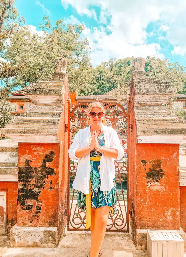 Em standing at the entrance of a temple in Lombok wearing a colorful dress and a light white cover-up, with hands in a respectful greeting pose.