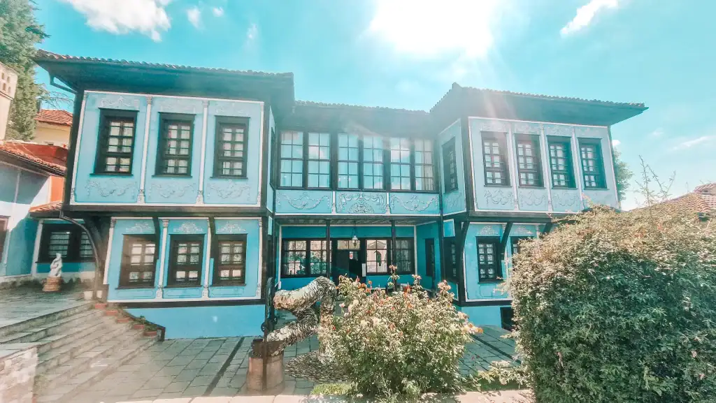 A light blue traditional Bulgarian Revival house in Plovdiv stands under the spring sun, its decorative wooden façade framed by flowering bushes and cobbled paths.