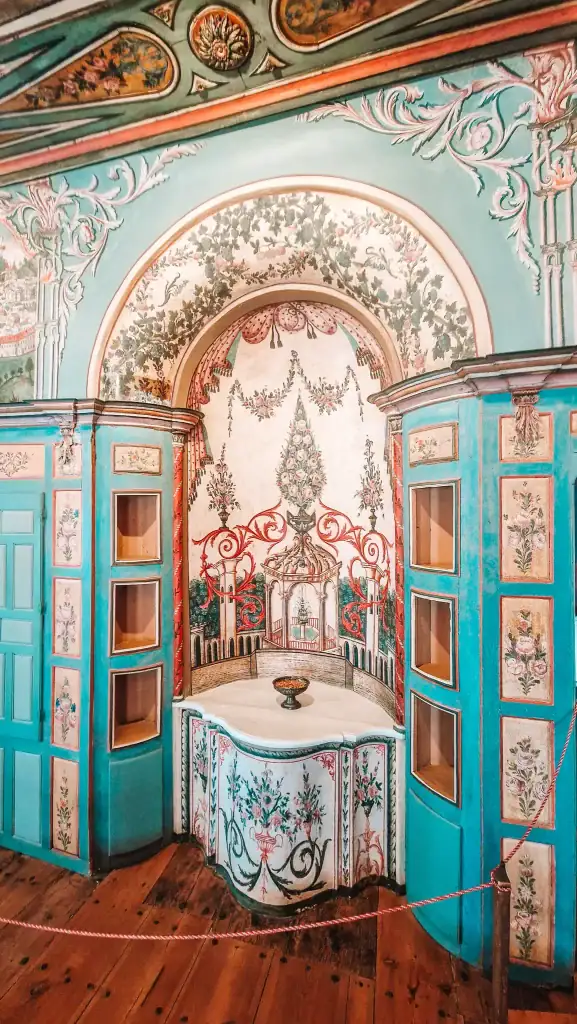 Ornately painted interior walls of a traditional Bulgarian home in Koprivshtitsa, featuring floral frescoes, turquoise woodwork, and a decorative built-in alcove with intricate detailing.