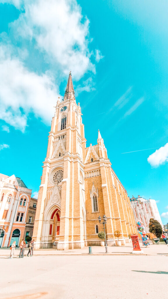 The towering yellow brick spire of the Name of Mary Church in Novi Sad, Serbia, rises into a vivid spring sky. Gothic-style architecture and a large rose window frame the entrance as visitors walk through the square below.