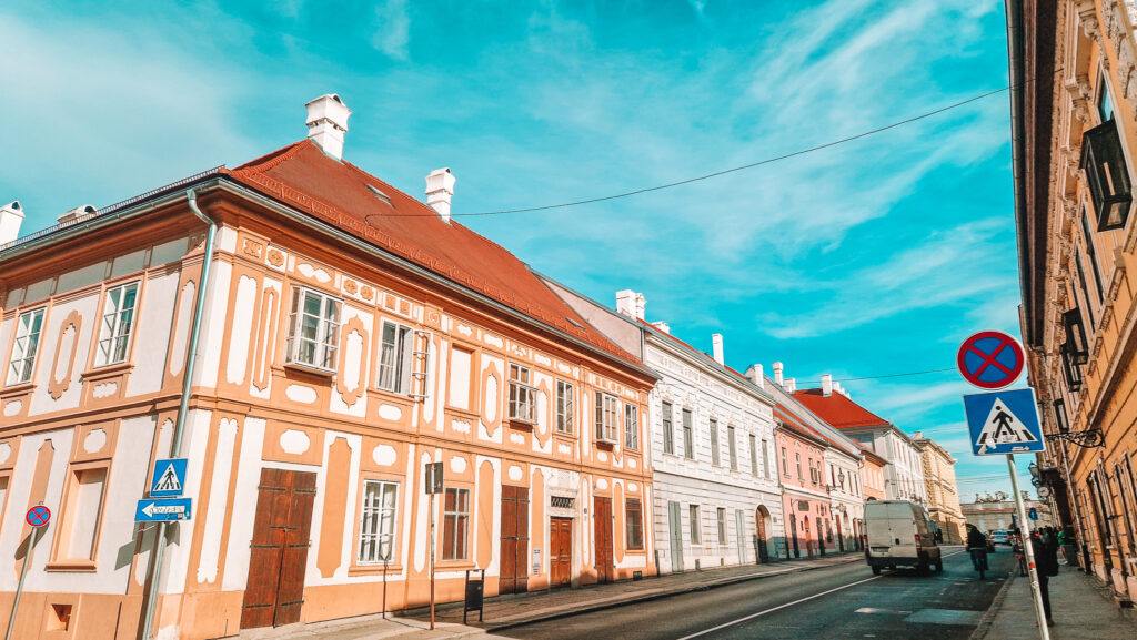 Ornately painted buildings with red roofs line a street in Novi Sad. The orange and cream Baroque-style architecture stands out against the bright spring sky, with pedestrians and cars passing by.