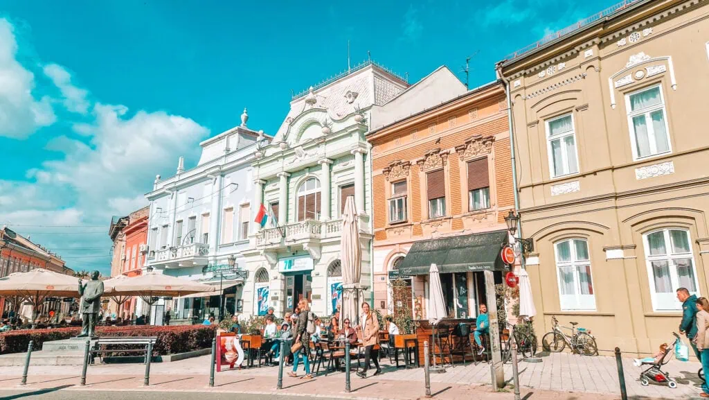 A lively pedestrian street in Novi Sad, Serbia, with locals enjoying outdoor cafes under parasols. Elegant pastel buildings with ornate facades line the street, and a statue of Jovan Jovanović Zmaj stands near the centre.