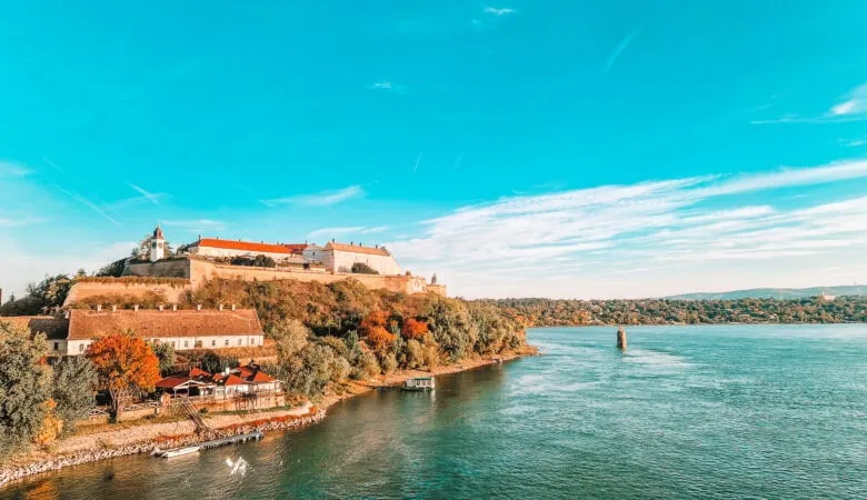 Petrovaradin Fortress in Novi Sad, Serbia, sits atop a tree-covered hill overlooking the Danube River. The red-roofed buildings and clock tower stand out against the clear blue sky, with a wide view of the riverbank.