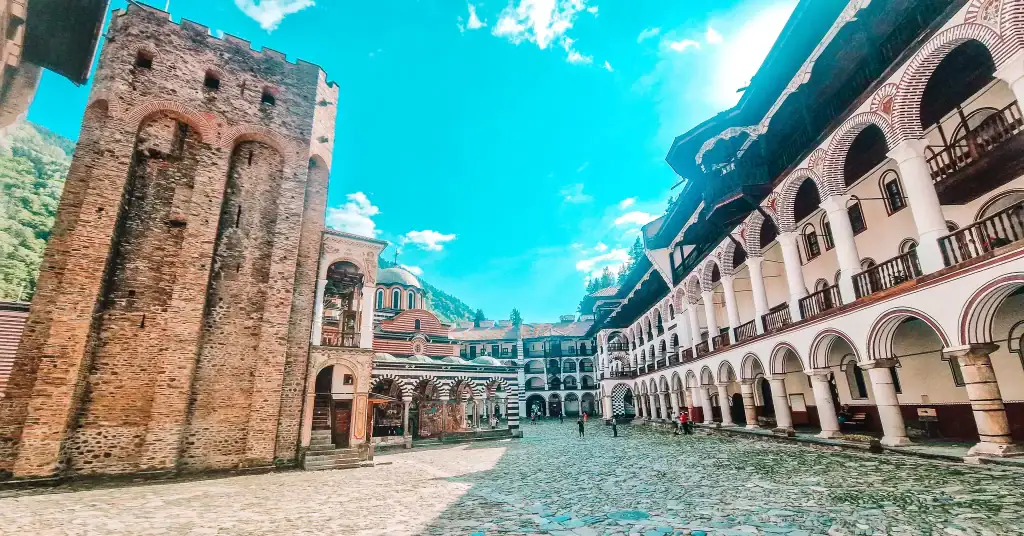 The courtyard of Rila Monastery in Bulgaria features a tall stone medieval tower, striped black-and-white arches, and multi-storey cloisters with wooden balconies, all backed by forested hills and a bright blue spring sky.