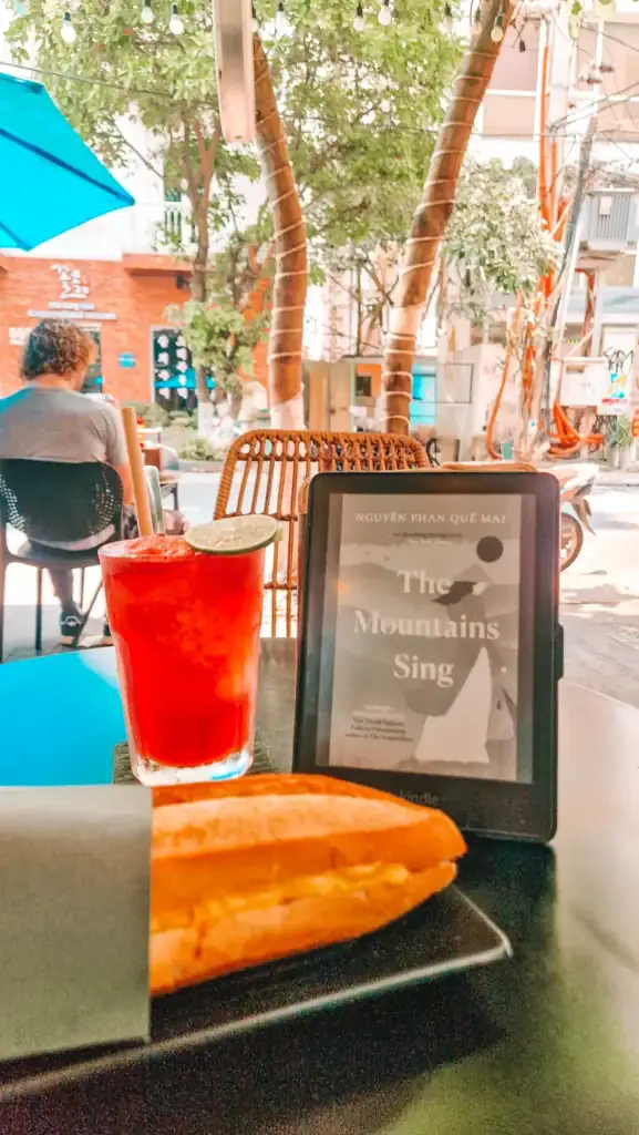 Kindle displaying the book The Mountains Sing next to a red drink with lime and a baguette-style sandwich at an outdoor café in Vietnam.
