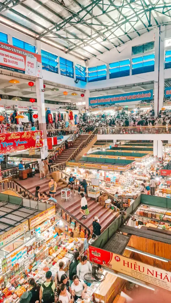 Bustling interior of Han Market in Da Nang with multiple levels of tightly packed stalls selling clothes, souvenirs, and snacks, filled with shoppers.