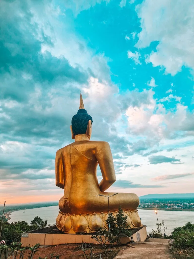 Back view of a large golden Buddha statue overlooking a wide river and a scenic valley at sunset, with dramatic clouds filling the sky.