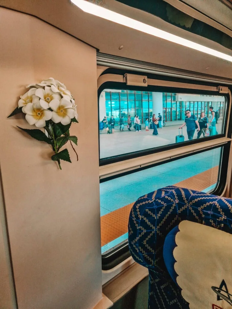 Inside a modern train carriage decorated with artificial white flowers, passengers outside are visible through the window at a busy station in Laos.