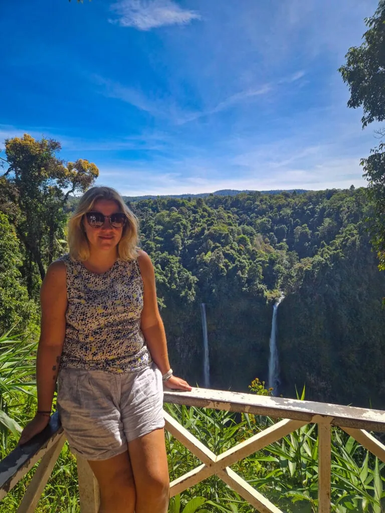 Em leans against a wooden railing overlooking a lush jungle valley with twin waterfalls cascading in the distance under a clear blue sky.